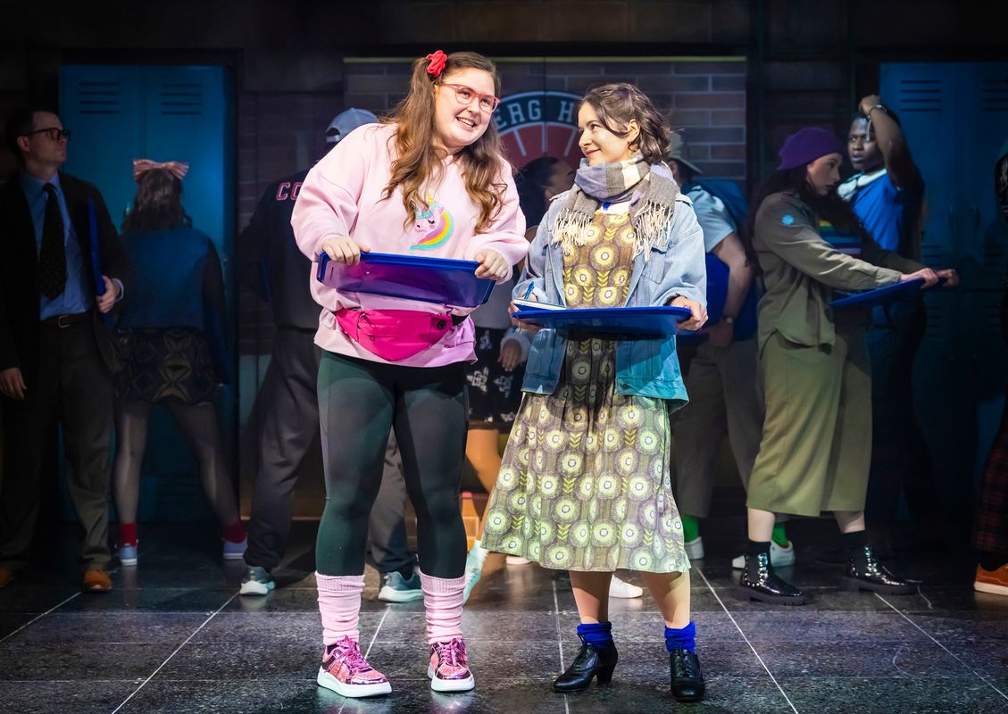 Two women on stage, smiling and holding blue trays, in front of school lockers. One wears a pink sweater, the other a patterned dress. Energetic mood.