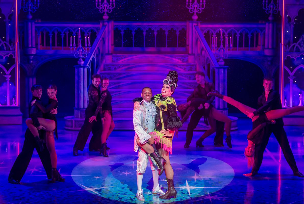 The wicked stepmother stands centre stage in a spotlight with their leg being held up by the prince.