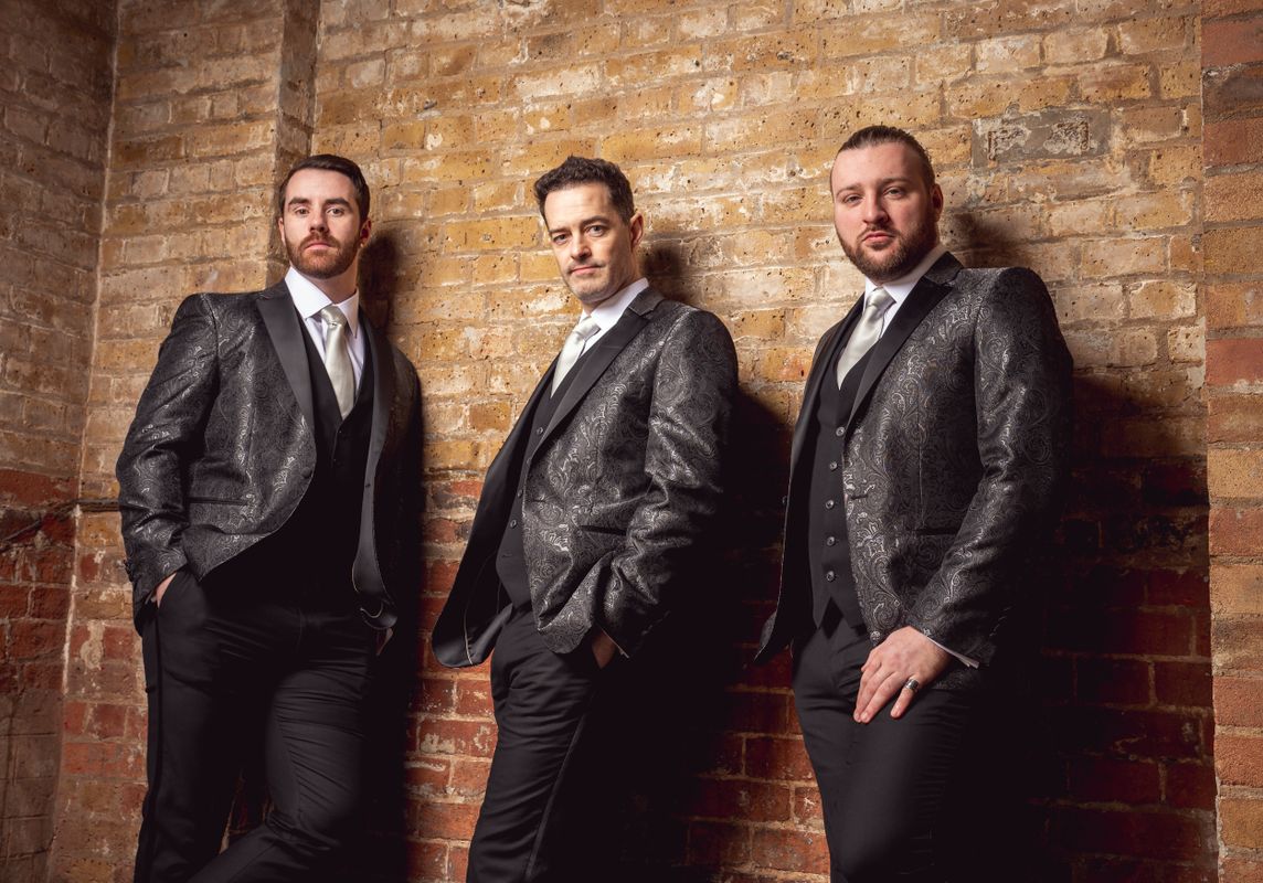 The Phantom boys in matching sparkly suits lean casually against a brick wall. They convey a confident and relaxed tone, wearing white shirts and ties.