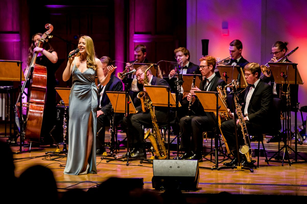 A singer wearing a long silver dressed is accompanied by the brass section.
