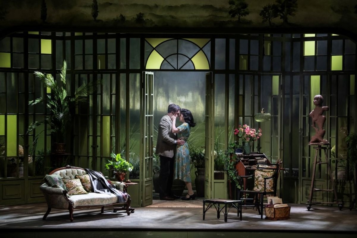 A theatrical stage set depicting an elegant conservatory with large windows and lush greenery. Two people are standing close together by the central door, surrounded by plants, a sofa, and a table with flowers and books. The scene has a romantic and vintage ambiance.