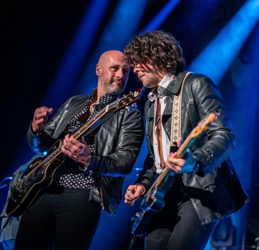 This photograph captures two members of The Illegal Eagles performing on stage, each playing electric guitar under dramatic blue lighting.