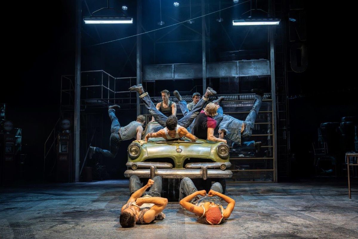 A dynamic stage scene with a performer in a red jacket and blue jeans energetically dancing on cars. A group of dancers energetically poses around, with dramatic lighting and an industrial backdrop creating an intense, lively atmosphere.