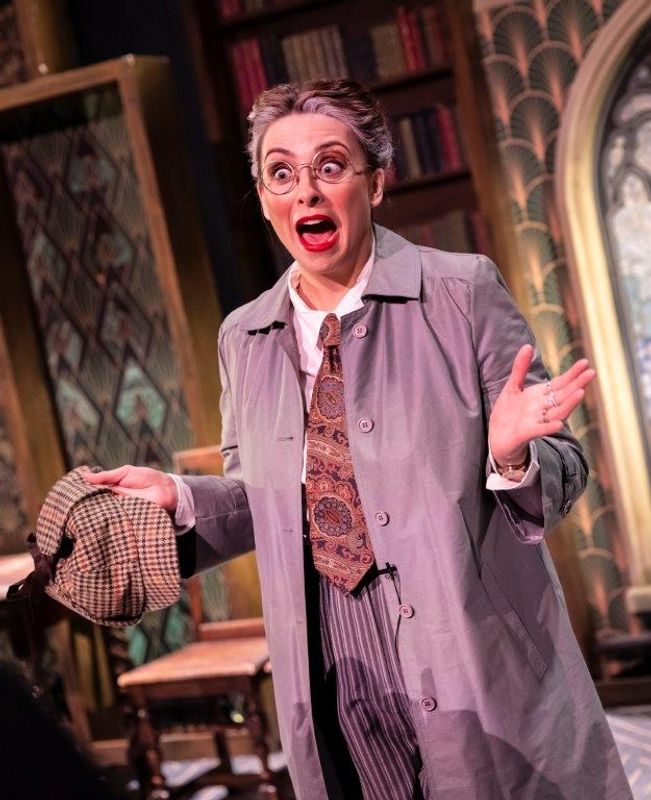 A performer in theatrical makeup and a grey suit gestures dramatically on stage. Background shows a bookcase and plants, creating a comedic tone.