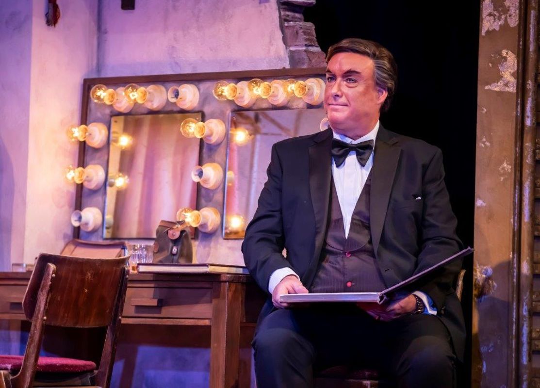 A man in a tuxedo sits holding a book backstage, beside a makeup mirror with warm, round lights. The scene conveys anticipation and focus.