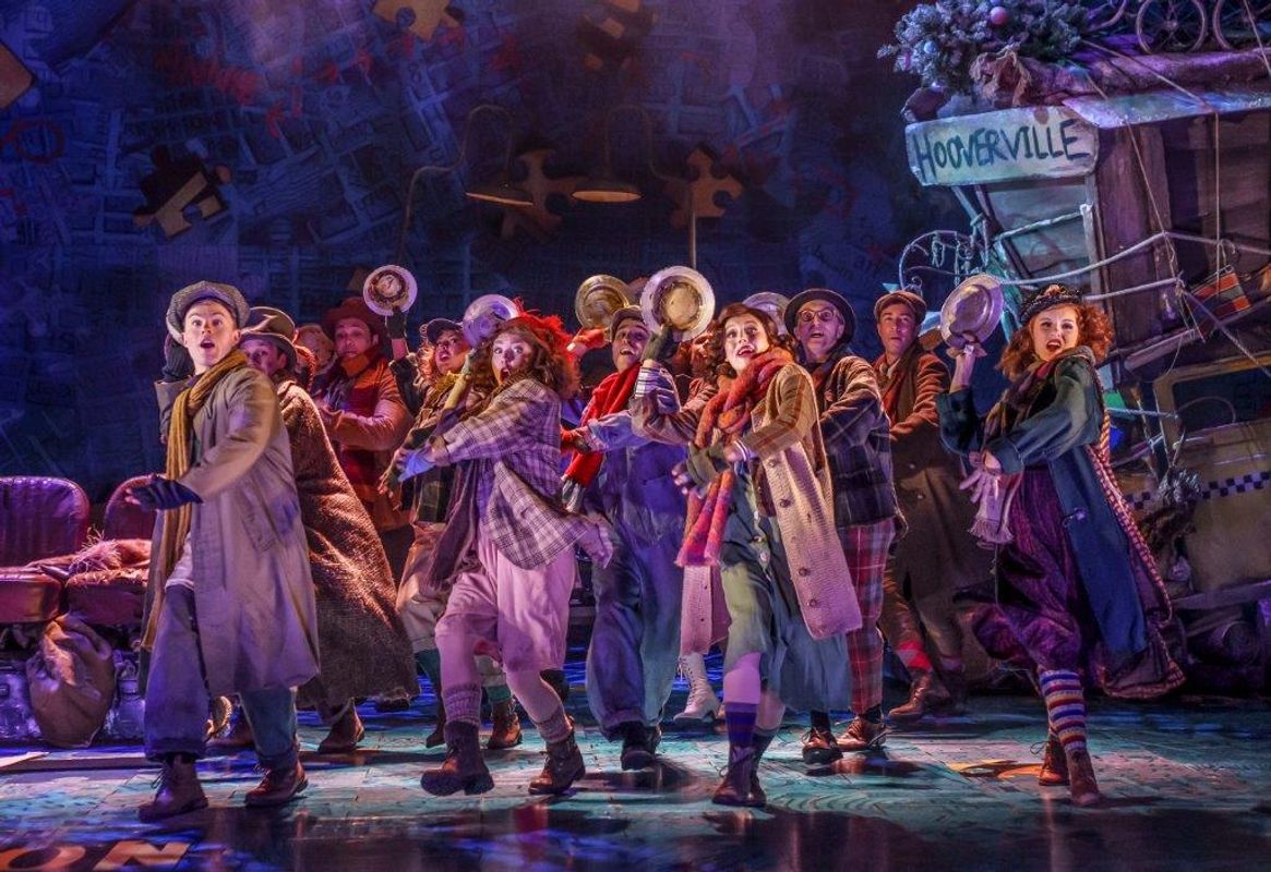 A lively theatrical scene with actors in vintage attire, holding hats, and showing dramatic expressions. A sign reads "Hooverville," suggesting a Depression-era setting.