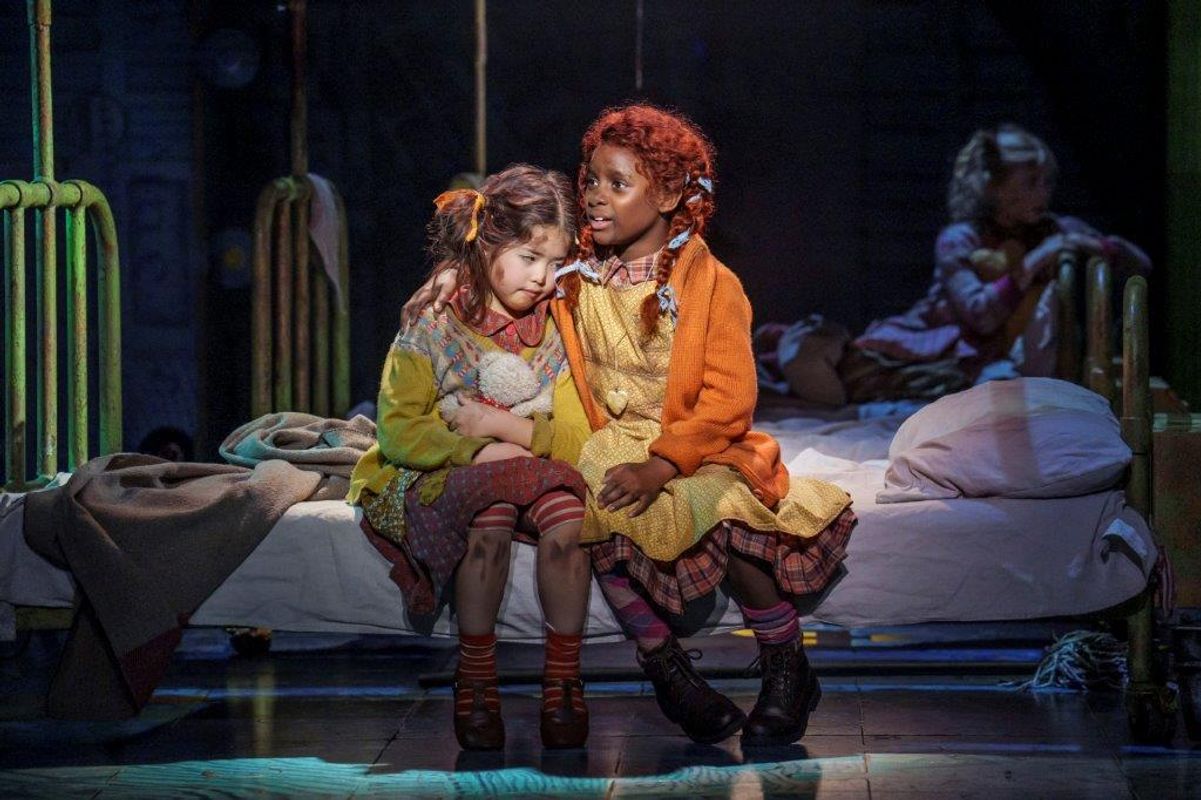 Two young girls in colorful outfits sit on a bed in a dimly lit room. One with red hair comforts the other. Another child is in the background.