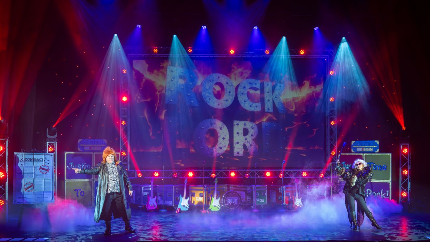 Two people stand on stage wearing elaborate clothing and wigs.