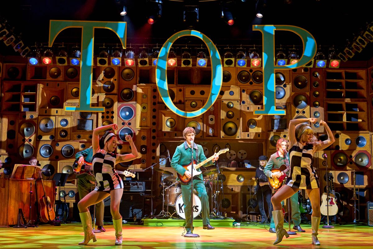 The band play on stage surrounded by dancers dressed in 1960 mini dresses.