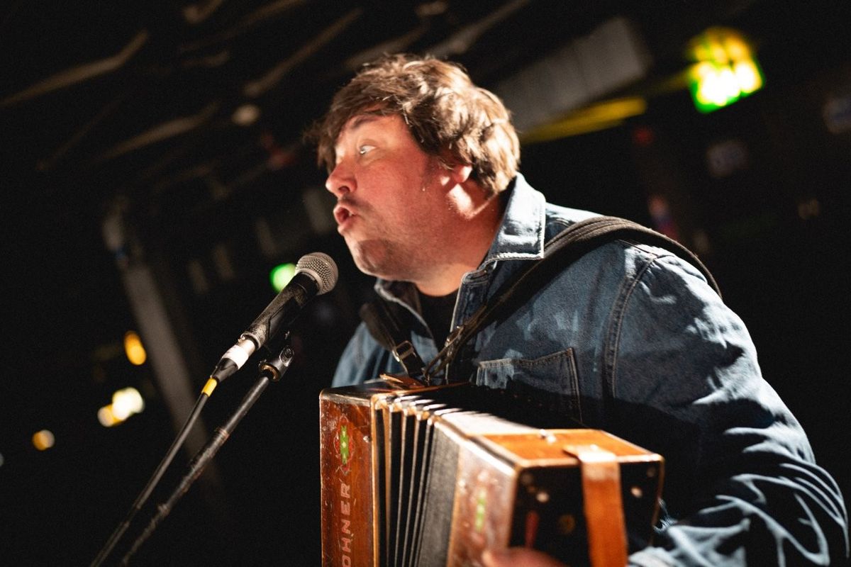 A musician performing on stage with an accordion, wearing a denim jacket and singing into a microphone. The intimate setting is lit by warm stage lights, creating a cozy atmosphere for the live performance.