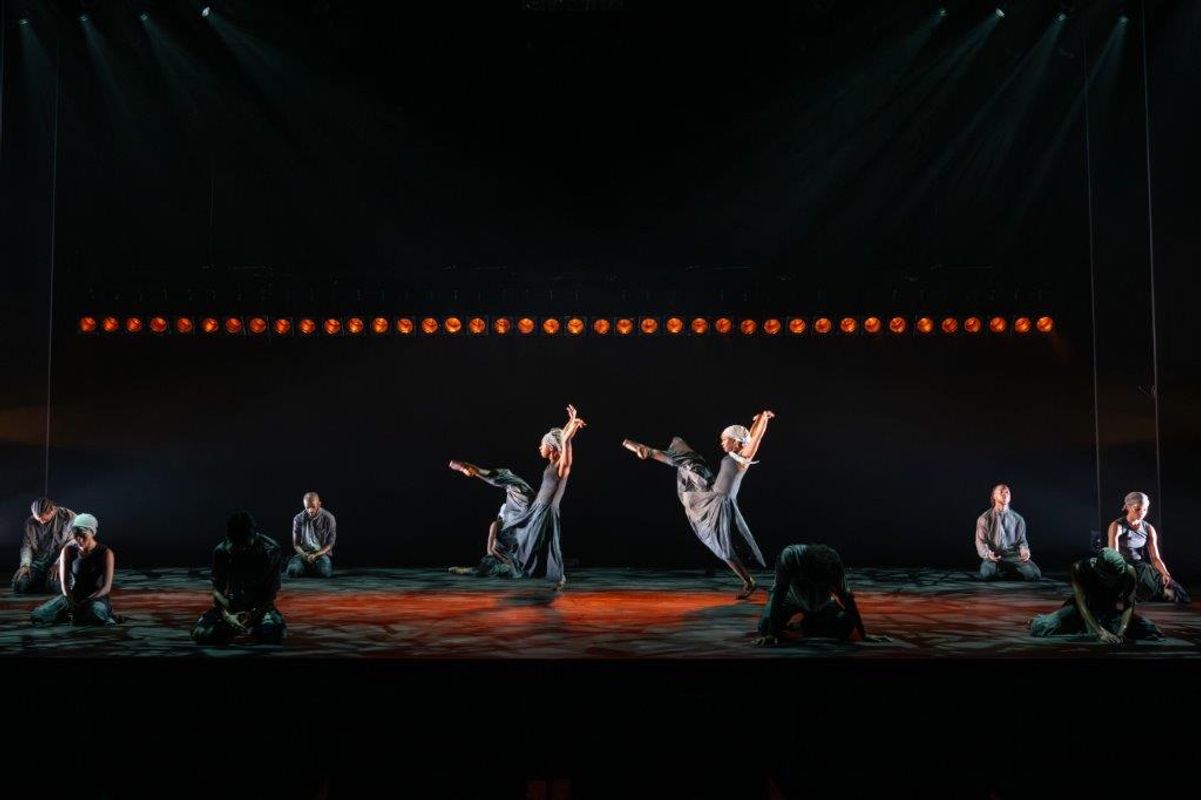 Group of dancers on stage wearing blue and grey outfits, a light casts a shadow over them, Two have their arms and a leg up in the air. others are kneeling around them