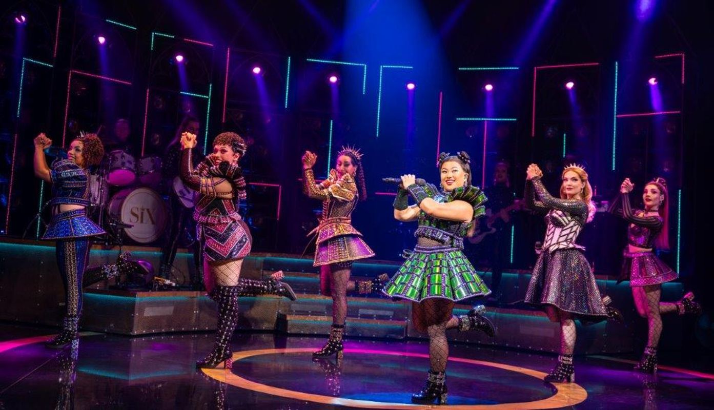 Six people stand on one leg clapping to their right hand side.