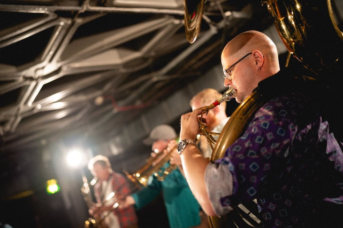 Four individuals are playing brass instruments in a dimly lit venue with a geometric ceiling pattern. The musicians appear focused, and a bright light source is visible in the background.