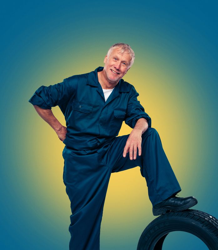 A person dressed in blue work overalls stands with their left leg raised on a black tyre.