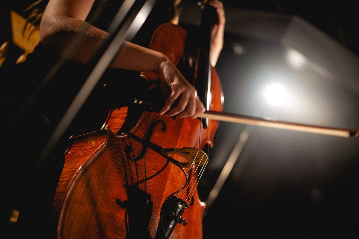 A person is playing a cello with a bow in a dimly lit environment. The focus is on the musician's hands and the instrument. The atmosphere is intimate and artistic.