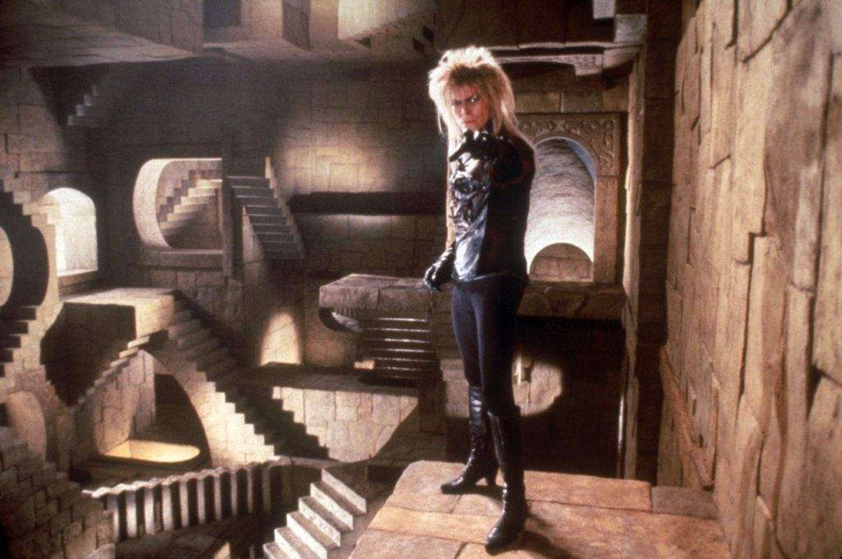 A person with choppy blond hair, wearing black heeled boots points to the camera.