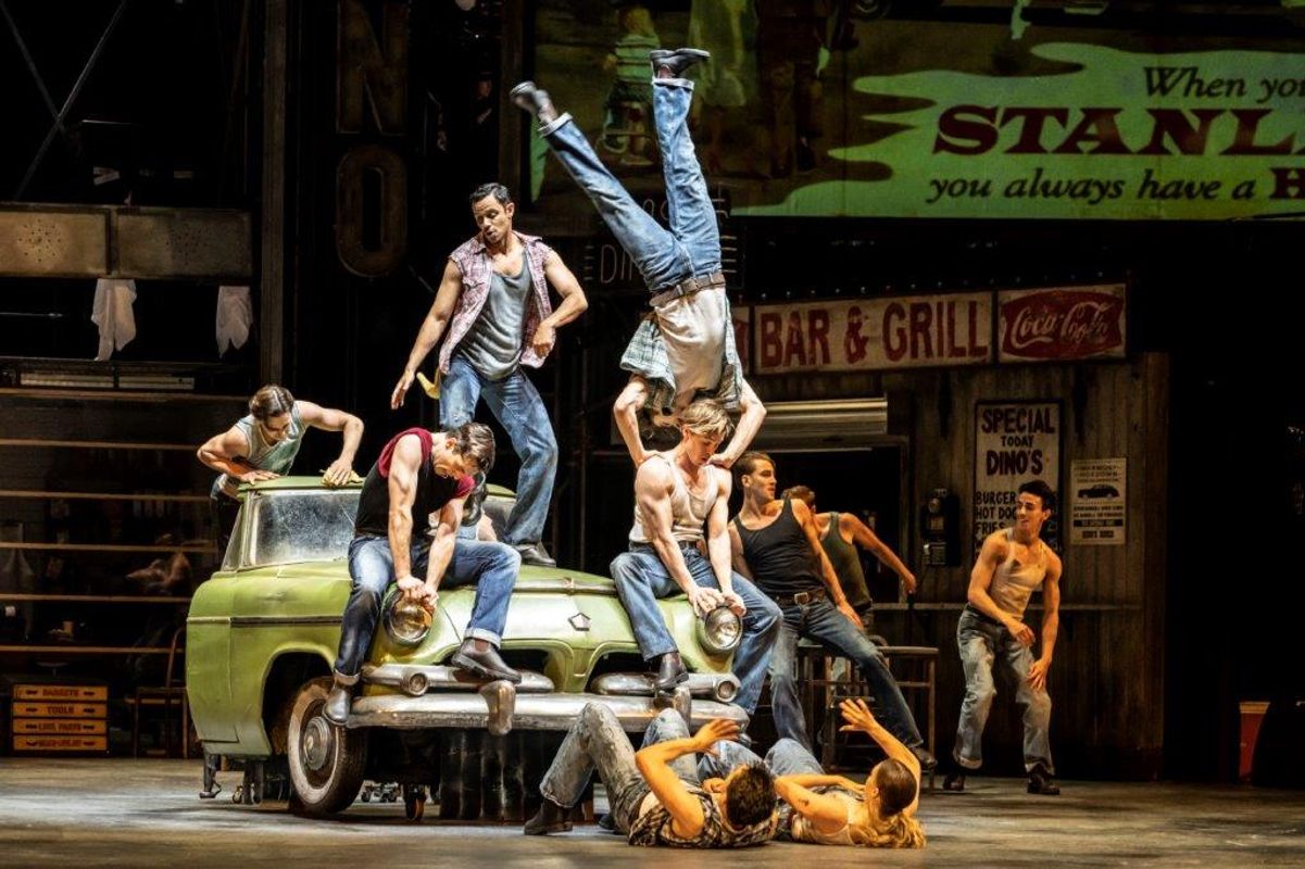 A dynamic dance scene onstage shows performers in casual attire, with one mid-flip on a vintage car. Nearby, others pose dramatically or lie on the ground.
