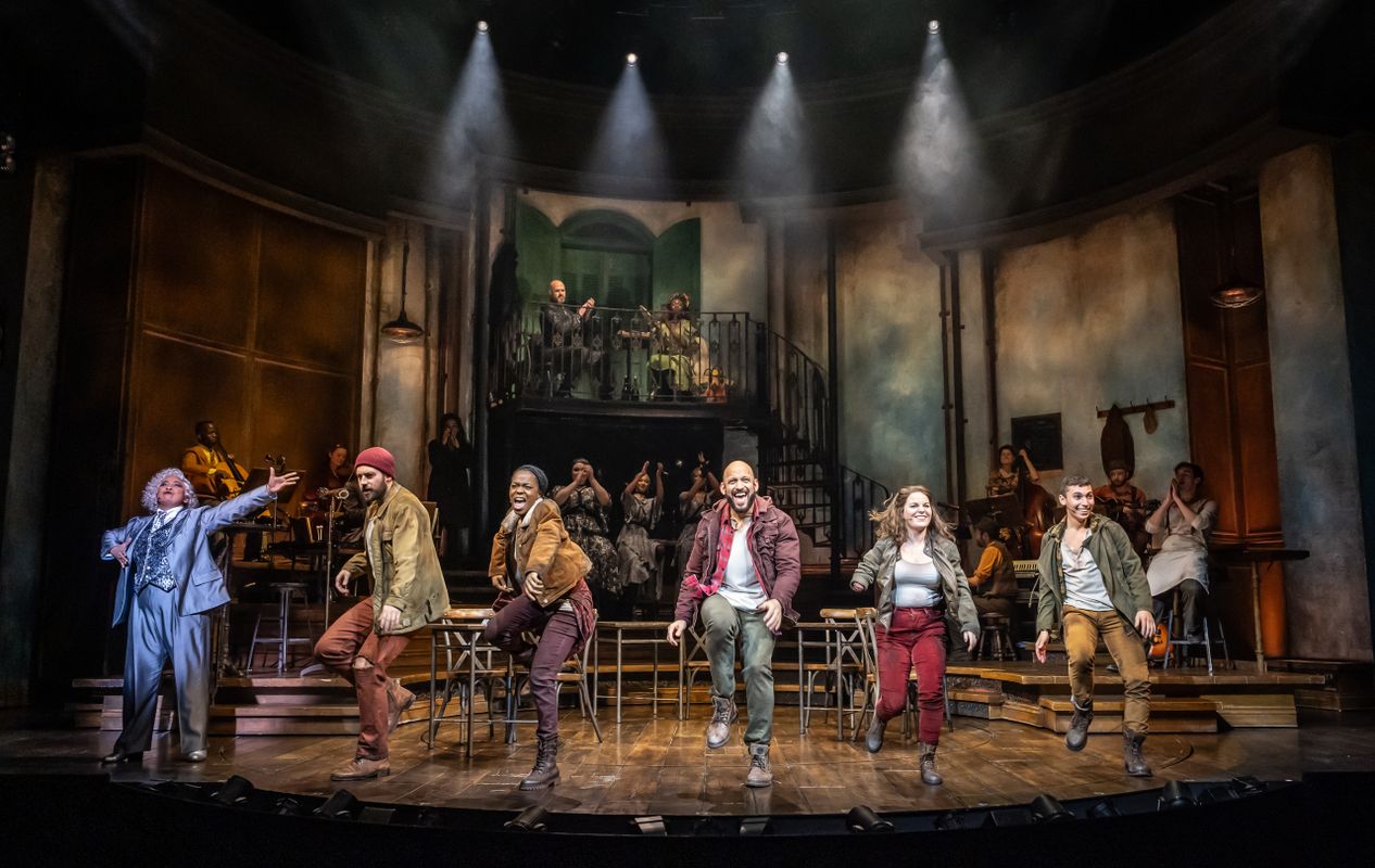 Six actors stand centre stage singing whilst the ensemble members in the background clap and cheer.