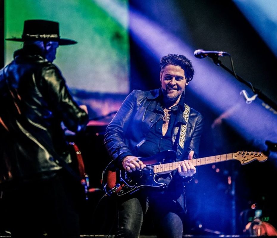 Two musicians are performing on stage. One is playing an electric guitar, wearing a dark leather jacket. The other is wearing a wide-brimmed hat and a leather jacket. The stage is illuminated with colorful lights.