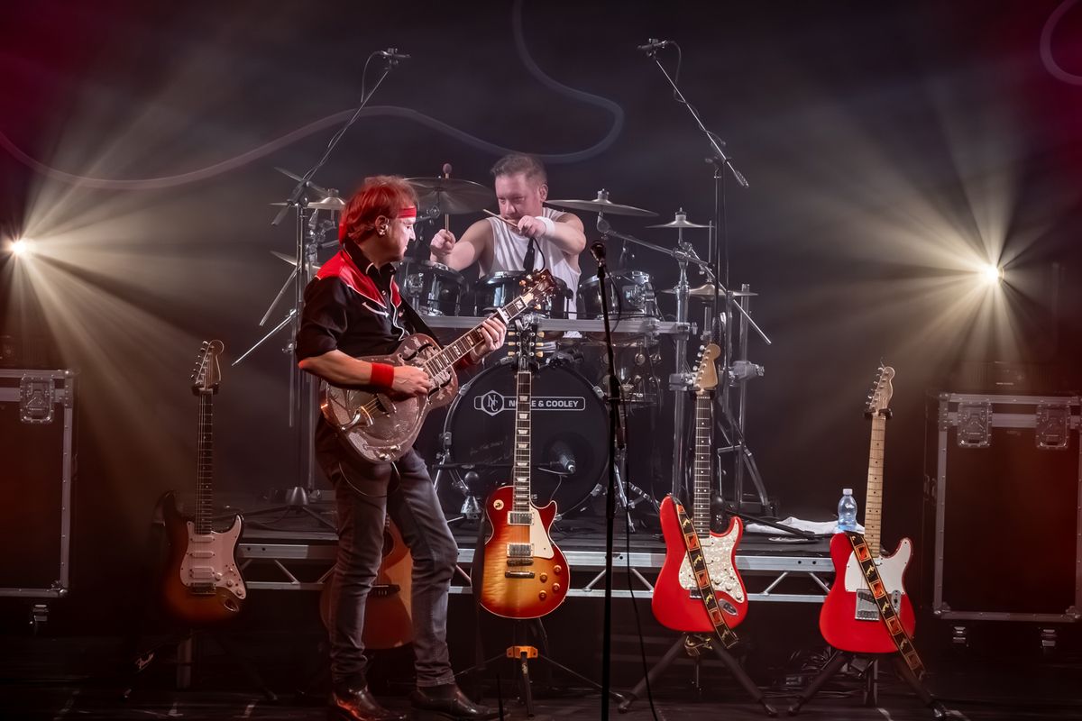 Band on stage performing, they're playing guitars and drums and there are flashing lights around them