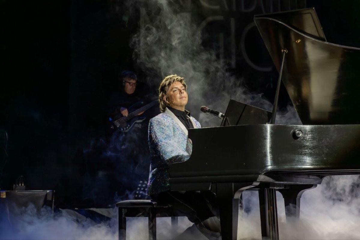 Martyn James in a sparkly jacket plays a grand piano on a dimly lit stage, surrounded by mist. A bassist is visible in the background, enhancing the moody atmosphere.
