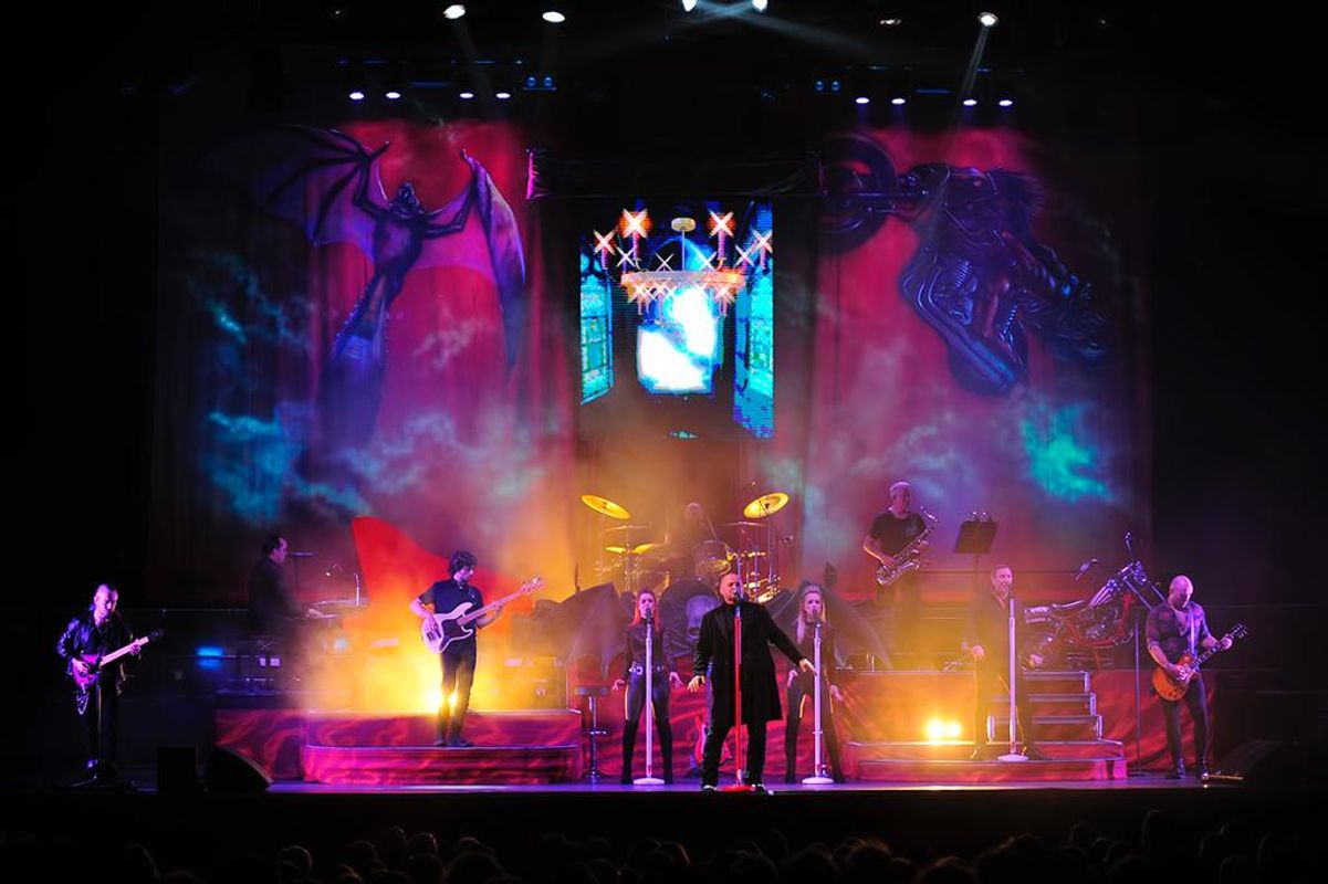 Steve Steinman and  band performing on stage with vibrant lights and dynamic backgrounds of a bat and mechanical horse. The scene is energetic and intense.