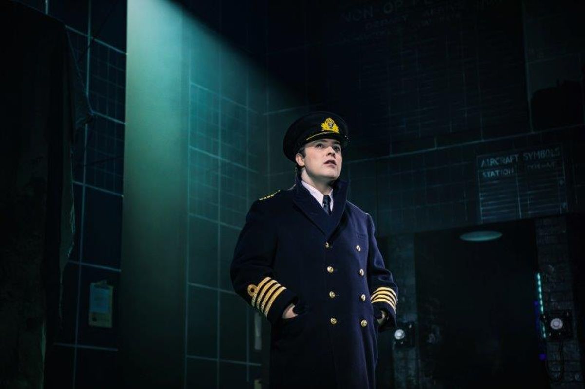 A pilot stands confidently under a spotlight in a dimly lit control room. He wears a navy uniform and cap, conveying authority and focus.