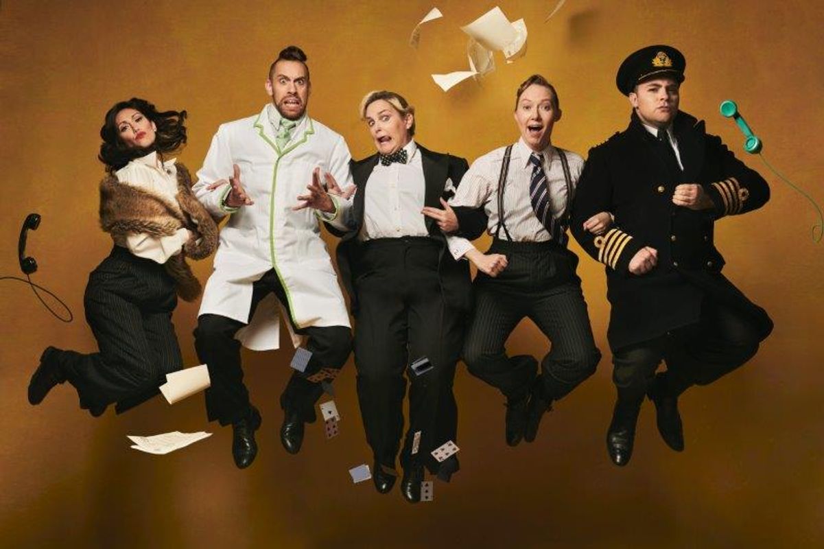 Five people in vintage attire jump against a brown backdrop, with papers and a phone floating mid-air. The scene conveys humor and chaos.