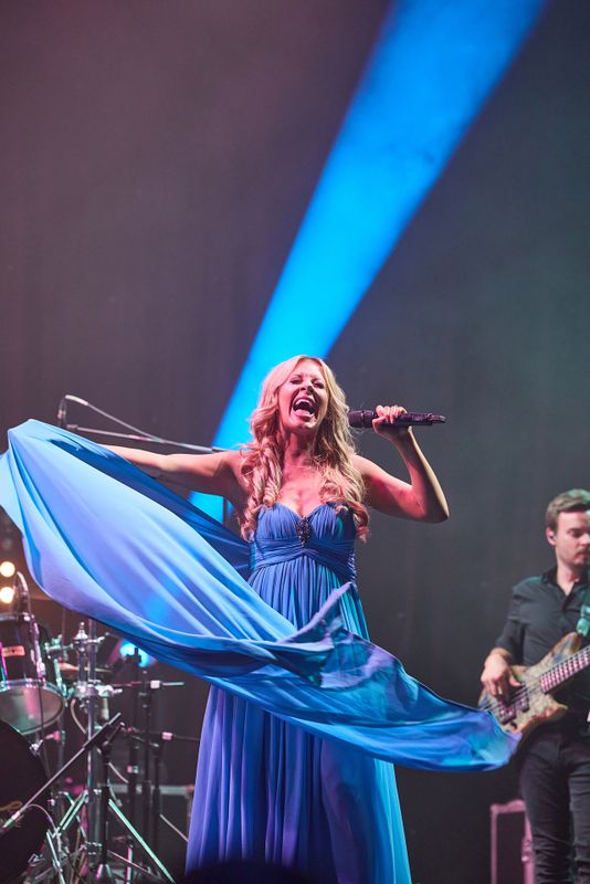 A performer in a flowing blue dress sings passionately on stage, holding a microphone. A bassist plays in the background, with bright stage lights and equipment visible.