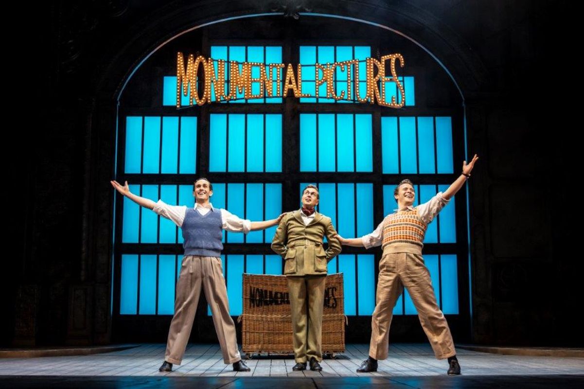 Three men stood in a line, the one in the middle has their arms folded behind him whilst the other two men have an arm stretched out
