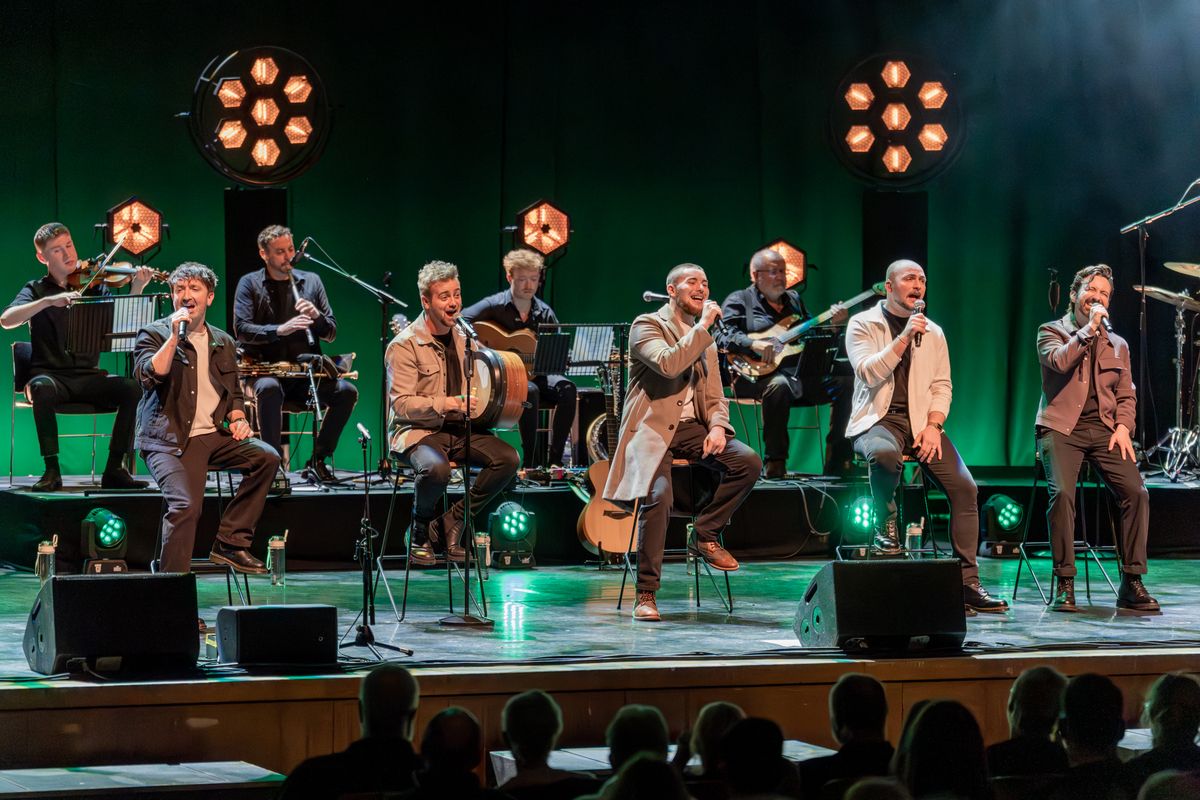 Five members are sat on tall stools singing into microphones. A band member sitting in one of the middle seats is playing a small drum