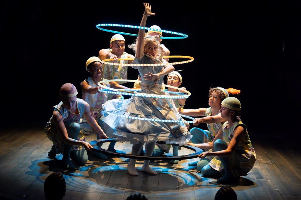 A woman is dancing in the centre of light up hoola hoops, there are dancers holding them up around her
