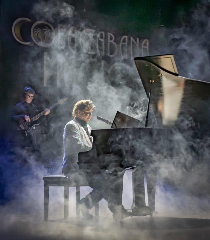 Martyn James in a sparkly jacket plays a grand piano on a dimly lit stage, surrounded by mist. A bassist is visible in the background, enhancing the moody atmosphere.