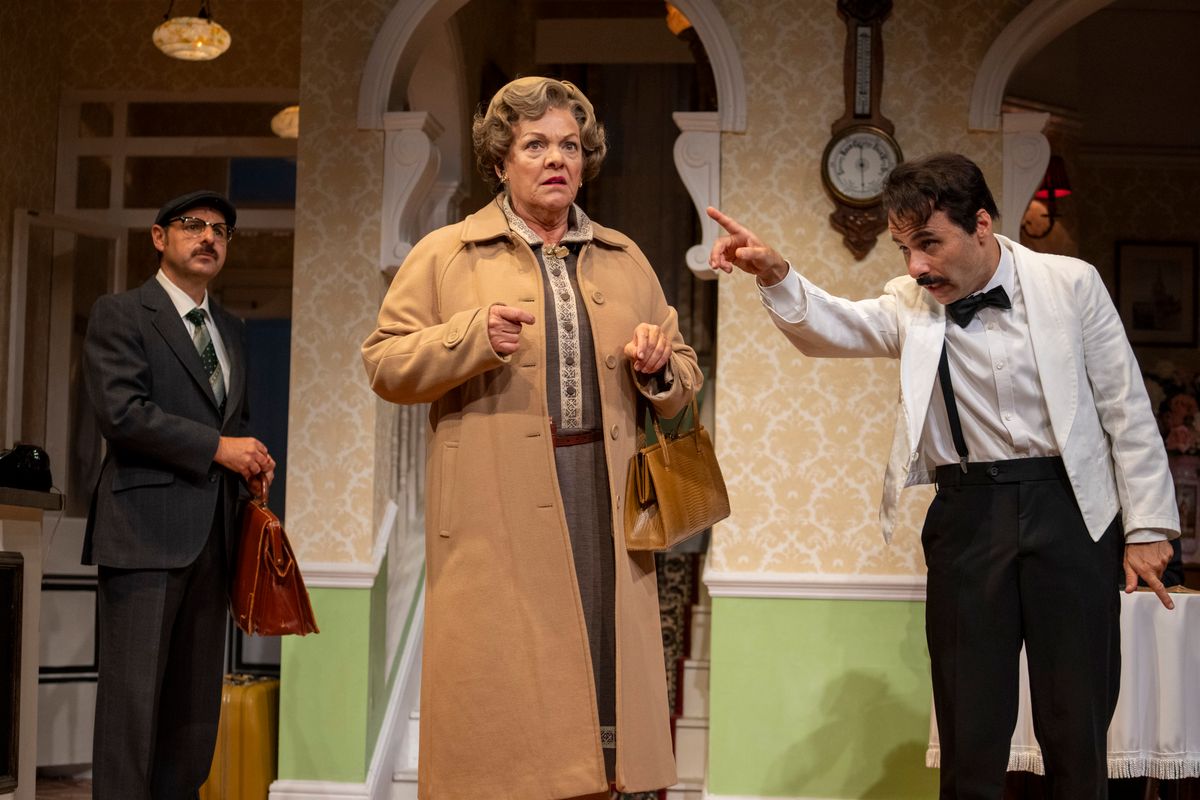 A person in a white jacket with a black moustache points at another person.