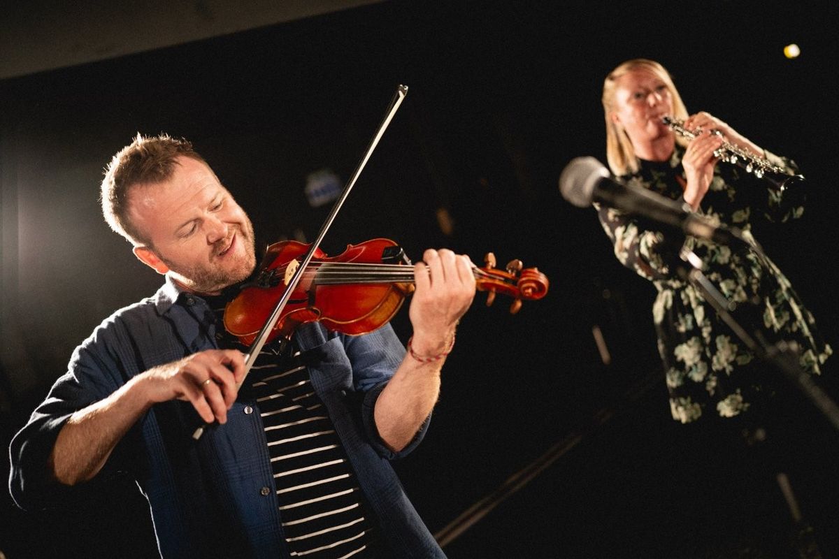 Two musicians performing on stage. One playing a violin and the other playing an oboe. Spotlights illuminate them, and a microphone is visible in the foreground.