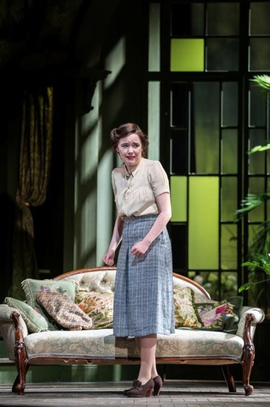 A woman wearing a light blouse and plaid skirt in a warmly lit set. She looks timid and worried.