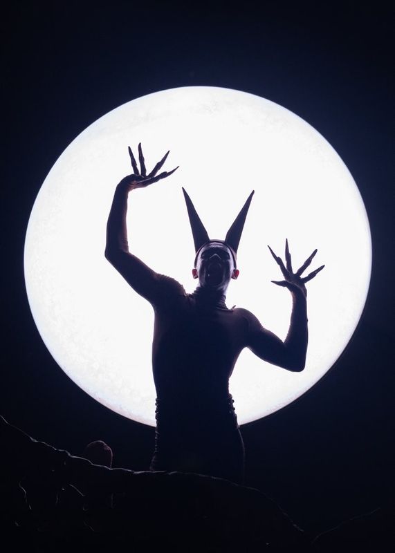 Silhouette of a person with claw-like hands and tall, pointed headgear, standing in front of a large, bright moon.