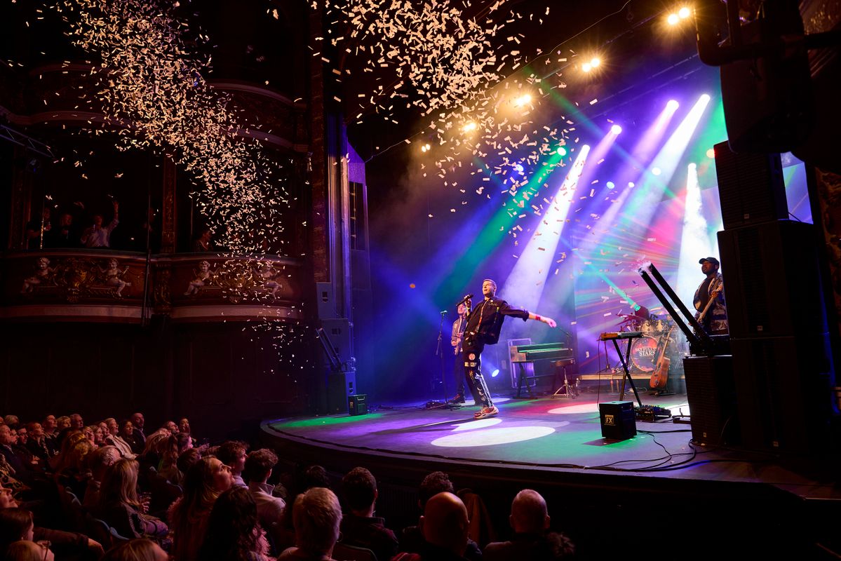 A performer energetically on stage with vibrant lights and confetti showering down, captivating a crowded audience in a lively theater setting.