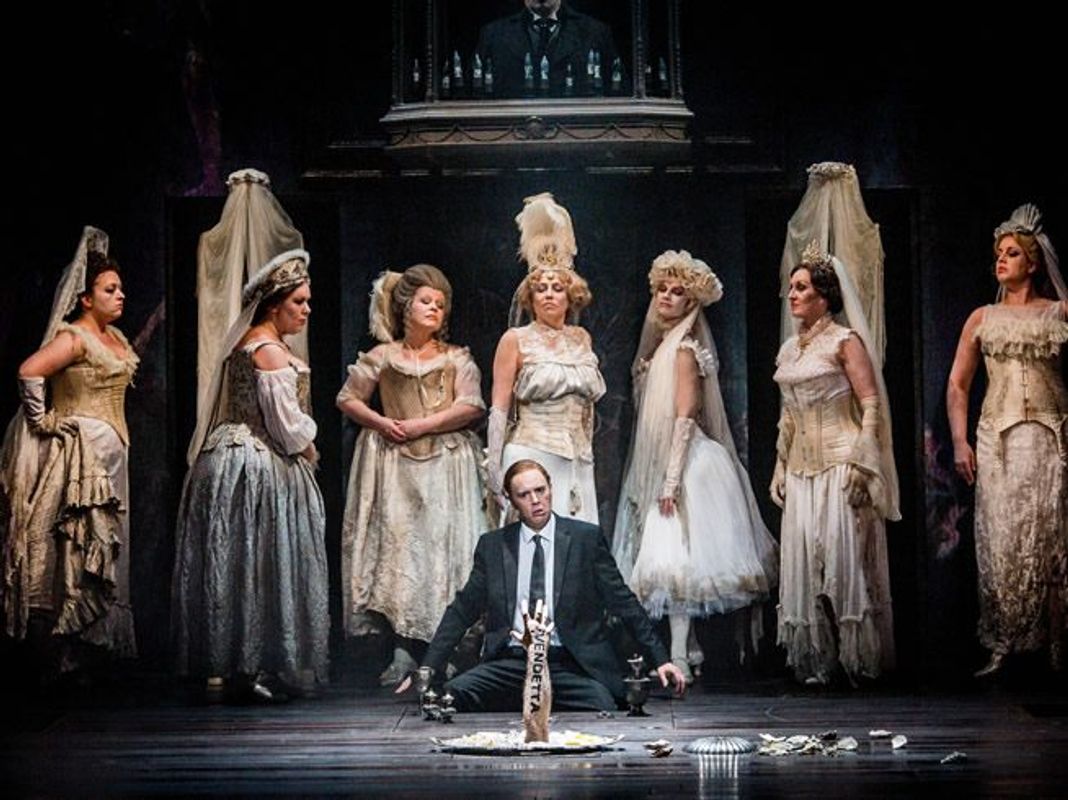 A person is sat on the floor infront of a silver tray. There is an arm coming out of the tray and it has the work 'Vendetta' written on it. There are a line of women stood behind the person, all in white Victorian dresses