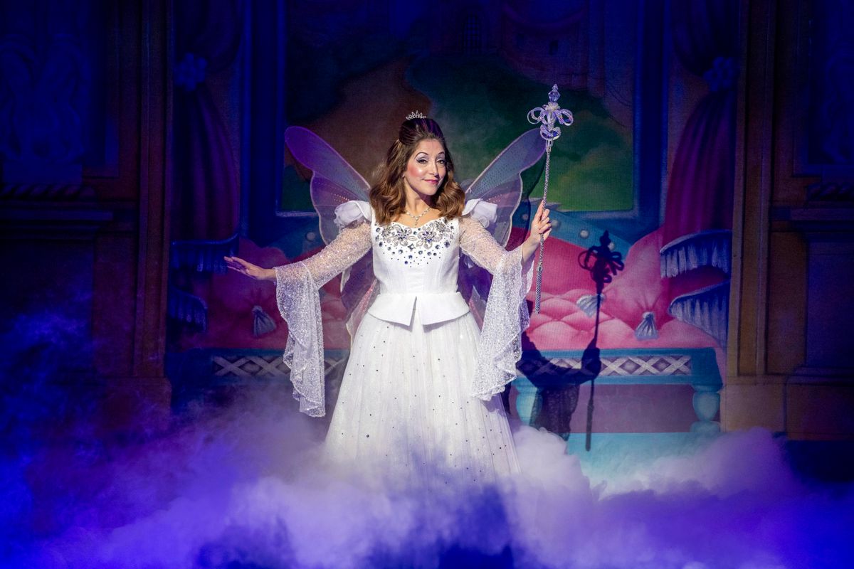 The Fairygodmother stands centre stage wearing a bejewelled white dress.