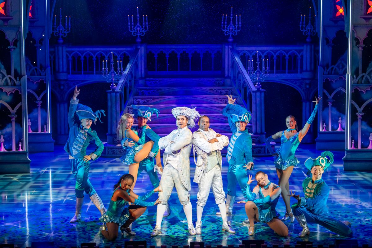 Prince charming and Dandini stand back to back dressed in white outfits with their arms folded.