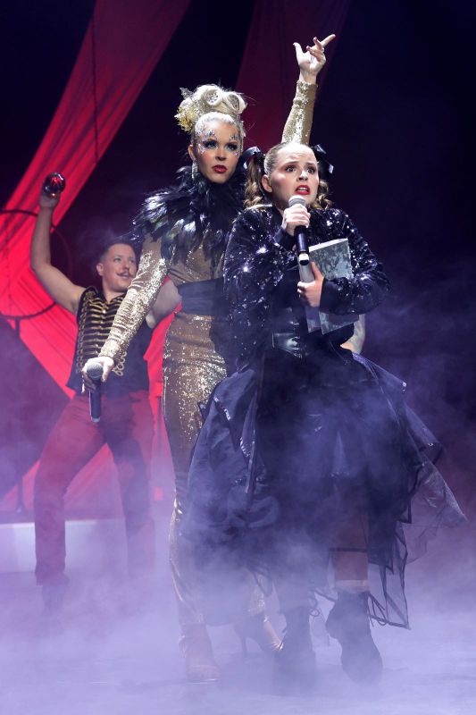 A person dressed in a black skirt and sequined jacket holds a book.