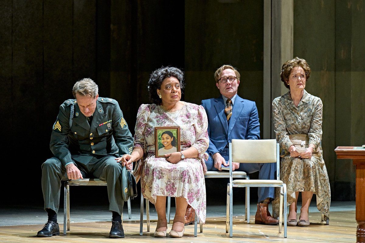 A general is sat on a seat with a woman who is holding a photo frame. There are two people sat behind them