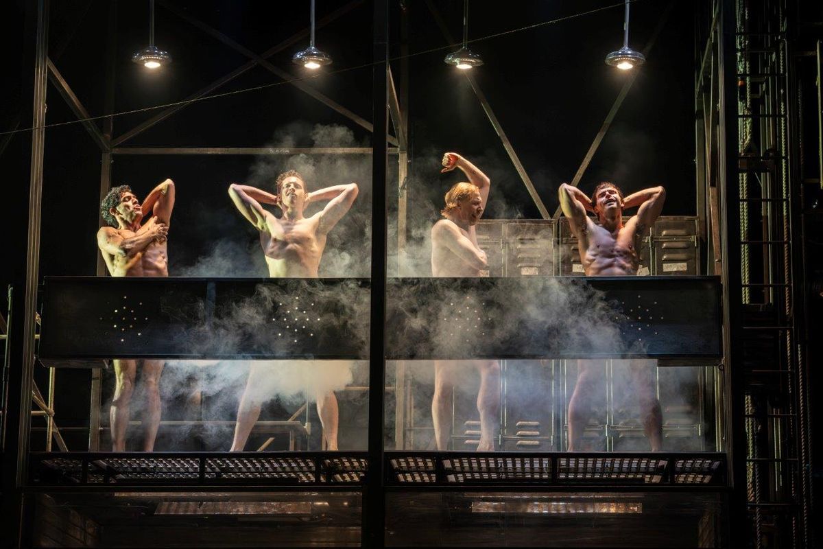 Four men showering on stage, surrounded by mist and dramatic lighting. They stand behind metal fixtures, creating an intense and artistic atmosphere.