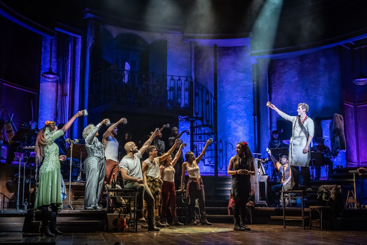 An actor stands centre stage with a red flower in their hair surrounded by ensemble members holding their tankards in the air to cheers.