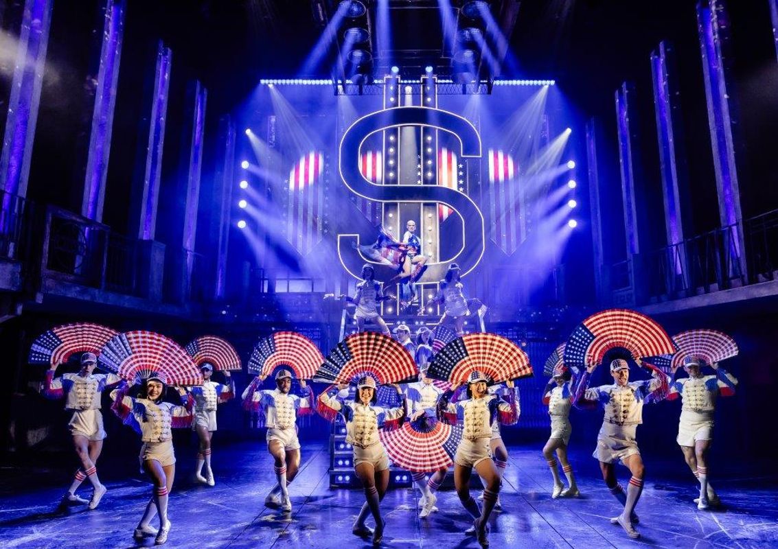 The ensemble dressed in white hold fans displaying the USA flag above their heads.