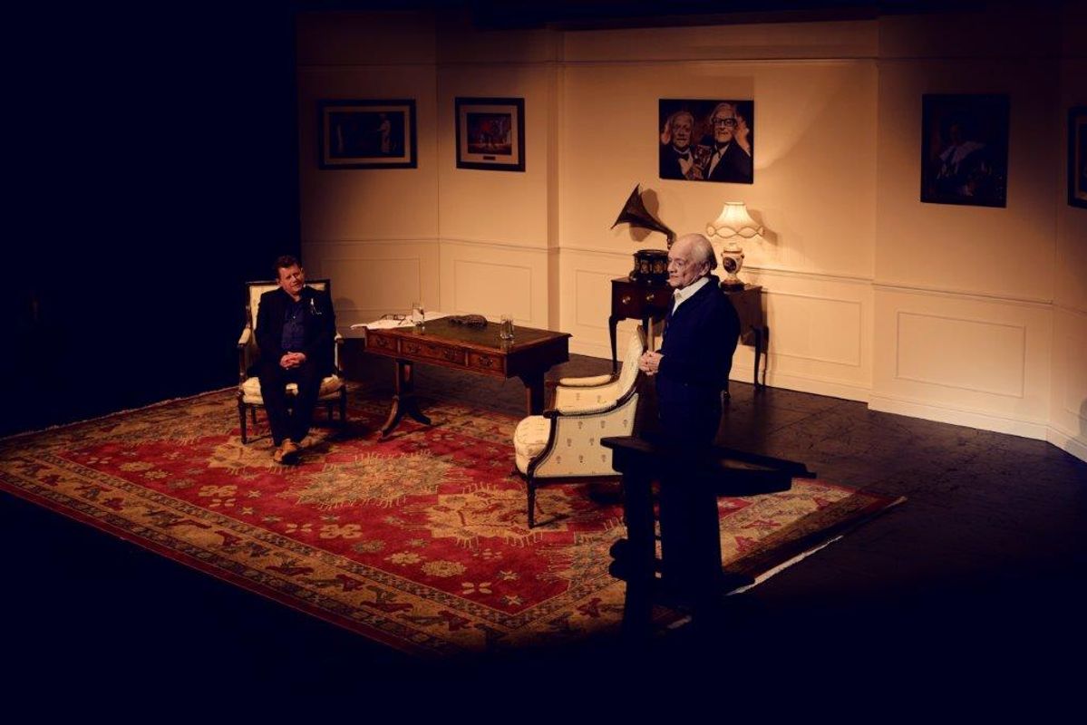 Sir David Jason on stage with another gentleman. The setting is made of two chairs, a dark wooden desk and a large rug