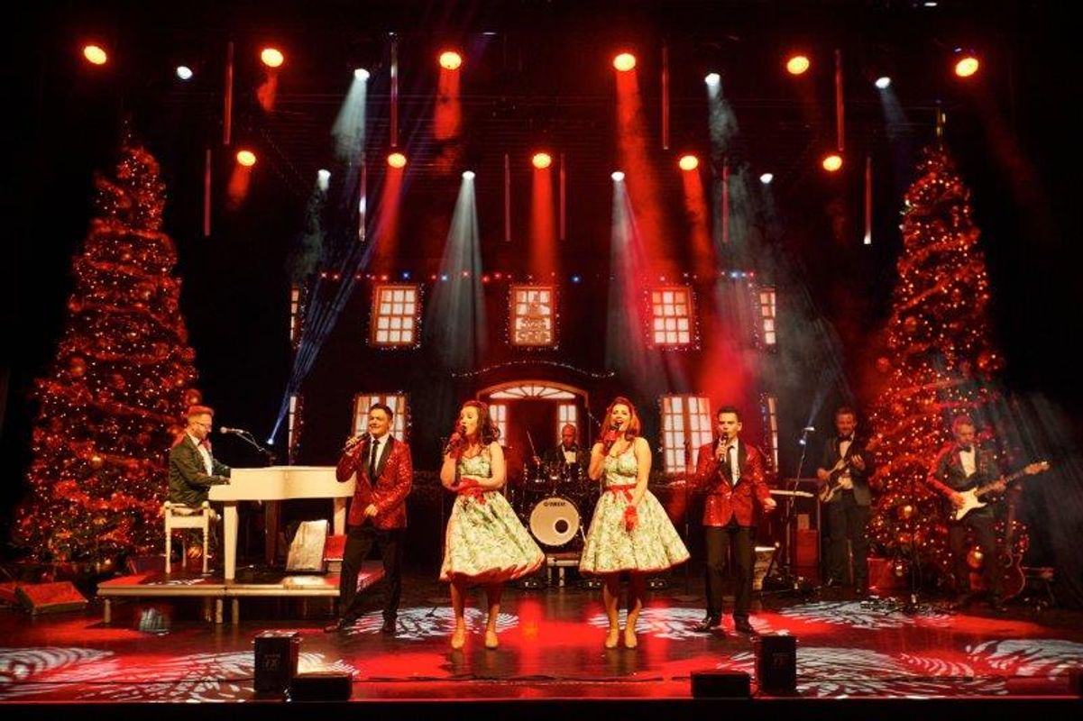 Four singers stand on stage surrounded by two Christmas trees.