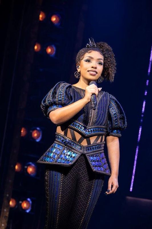 A person wearing a black and blue studded outfit smiles on stage.