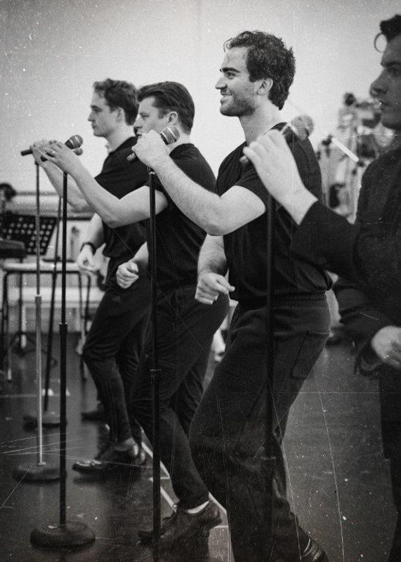 Black and white image of cats members rehearsing for Jersey boys. They are all wearing black and in a rehearsal studio